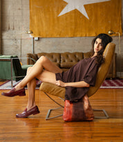 A person wearing dark red leather loafers sits in a leather chair with a multicolored Bed|Stü Dallas LTC leather shoulder bag at their feet.