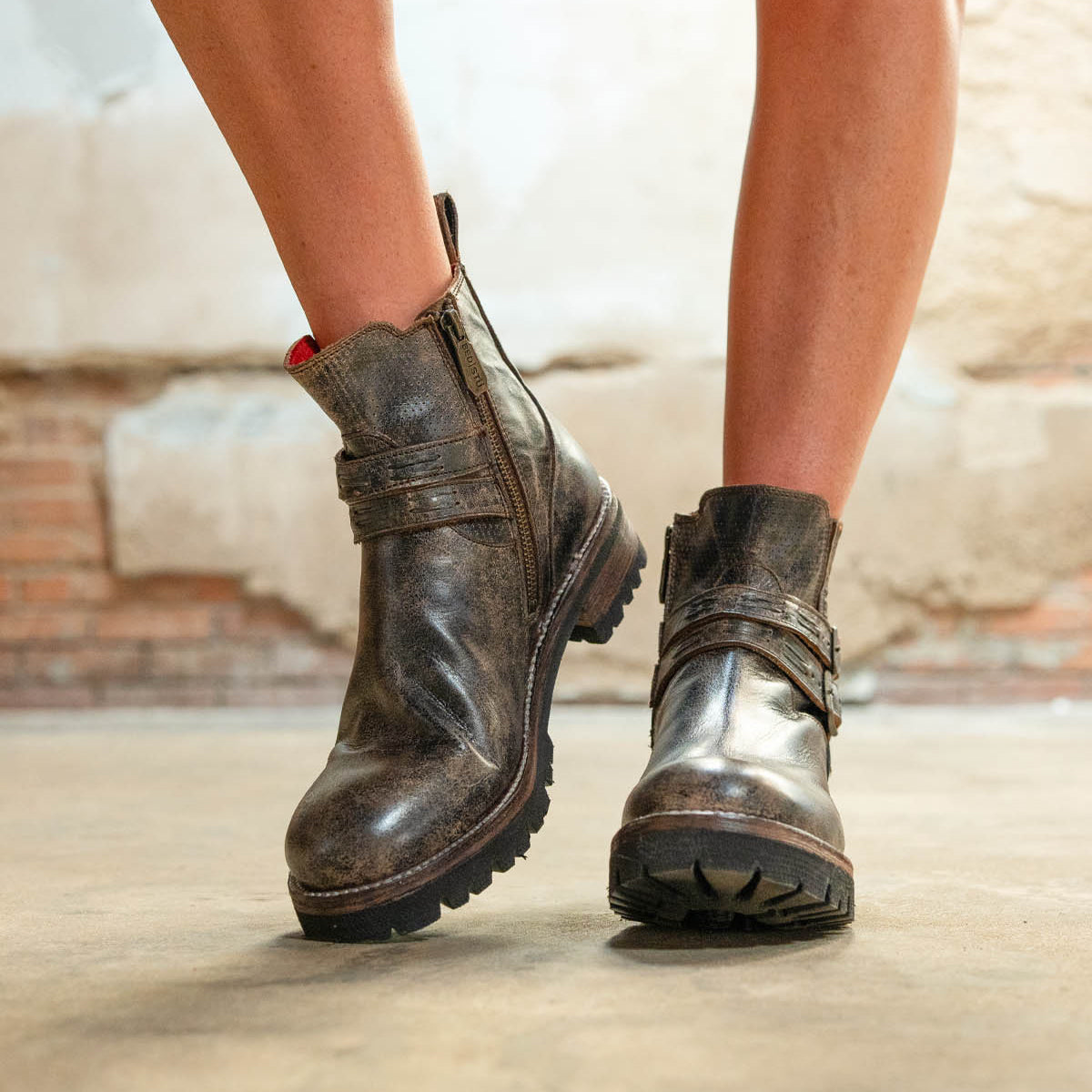 A person stands on a concrete floor wearing Bed Stu Daring boots—worn brown leather ankle boots with zippers, adjustable buckles, and a stacked heel.