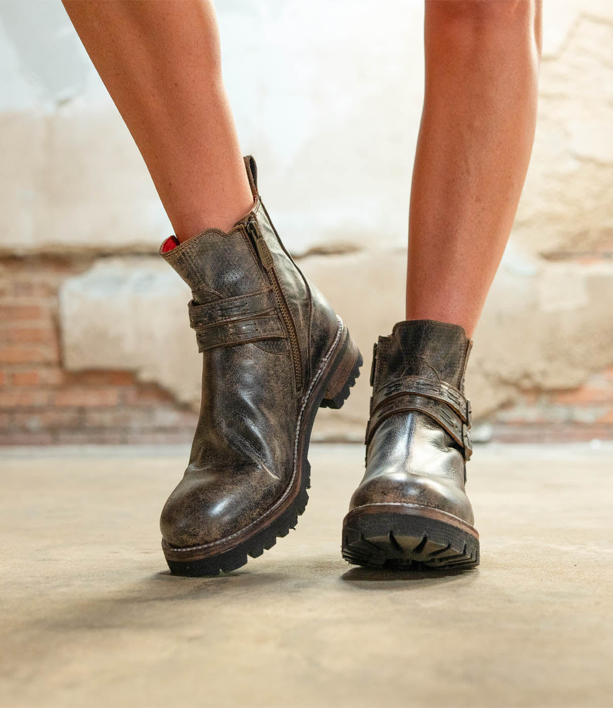 A person stands on a concrete floor wearing Bed Stu Daring boots—worn brown leather ankle boots with zippers, adjustable buckles, and a stacked heel.