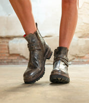 A person stands on a concrete floor wearing Bed Stu Daring boots—worn brown leather ankle boots with zippers, adjustable buckles, and a stacked heel.