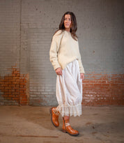 A woman stands indoors against a brick wall, wearing a cream sweater, long white skirt with lace trim, and Bed Stu's Daring ankle boots featuring adjustable buckle straps.
