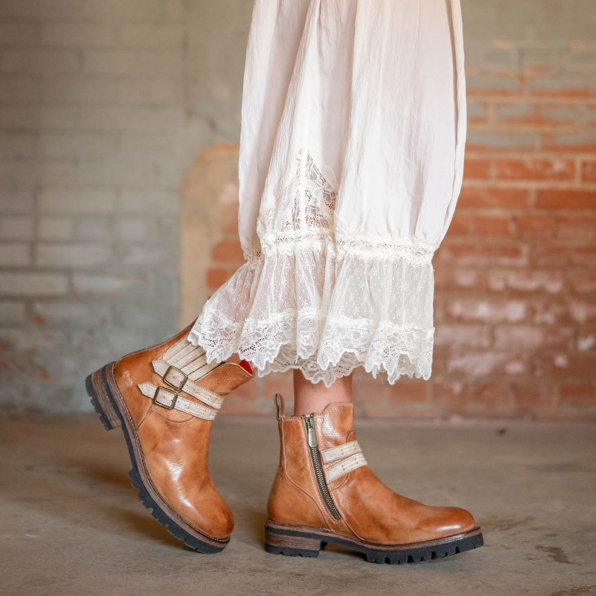 Wearing the Daring tan leather ankle boots by Bed Stu, featuring adjustable buckle straps, a person stands against a textured brick wall in a long white lace-trimmed skirt.