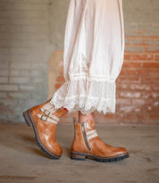 Wearing the Daring tan leather ankle boots by Bed Stu, featuring adjustable buckle straps, a person stands against a textured brick wall in a long white lace-trimmed skirt.