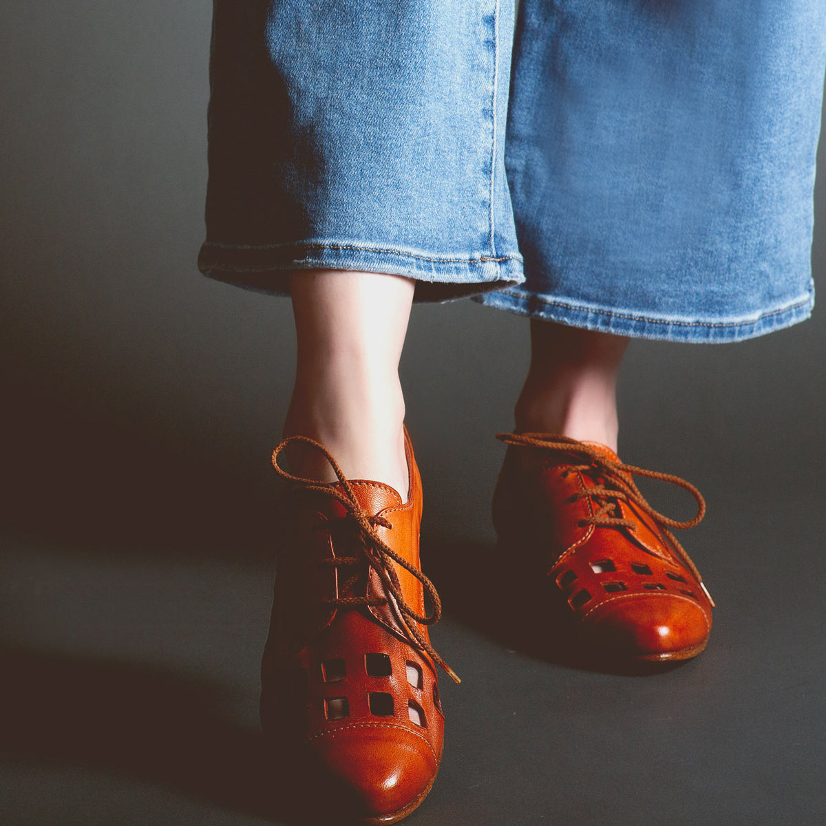 Person wearing wide-leg blue jeans and unique women's footwear in the form of Decoy by Bed Stu, brown leather oxford shoes with cut-out details, standing on a dark surface.