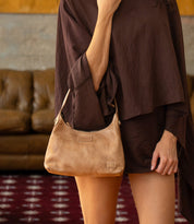 A person wearing a brown tunic holds a Bed|Stü Denton, a small, light tan vegetable-tanned leather shoulder bag with a strap.