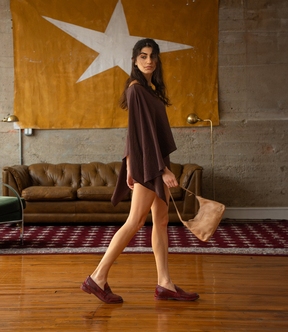 A woman walks wearing dark red leather loafers, a brown poncho, and carrying a vegetable-tanned leather Denton shoulder bag by Bed|Stü.