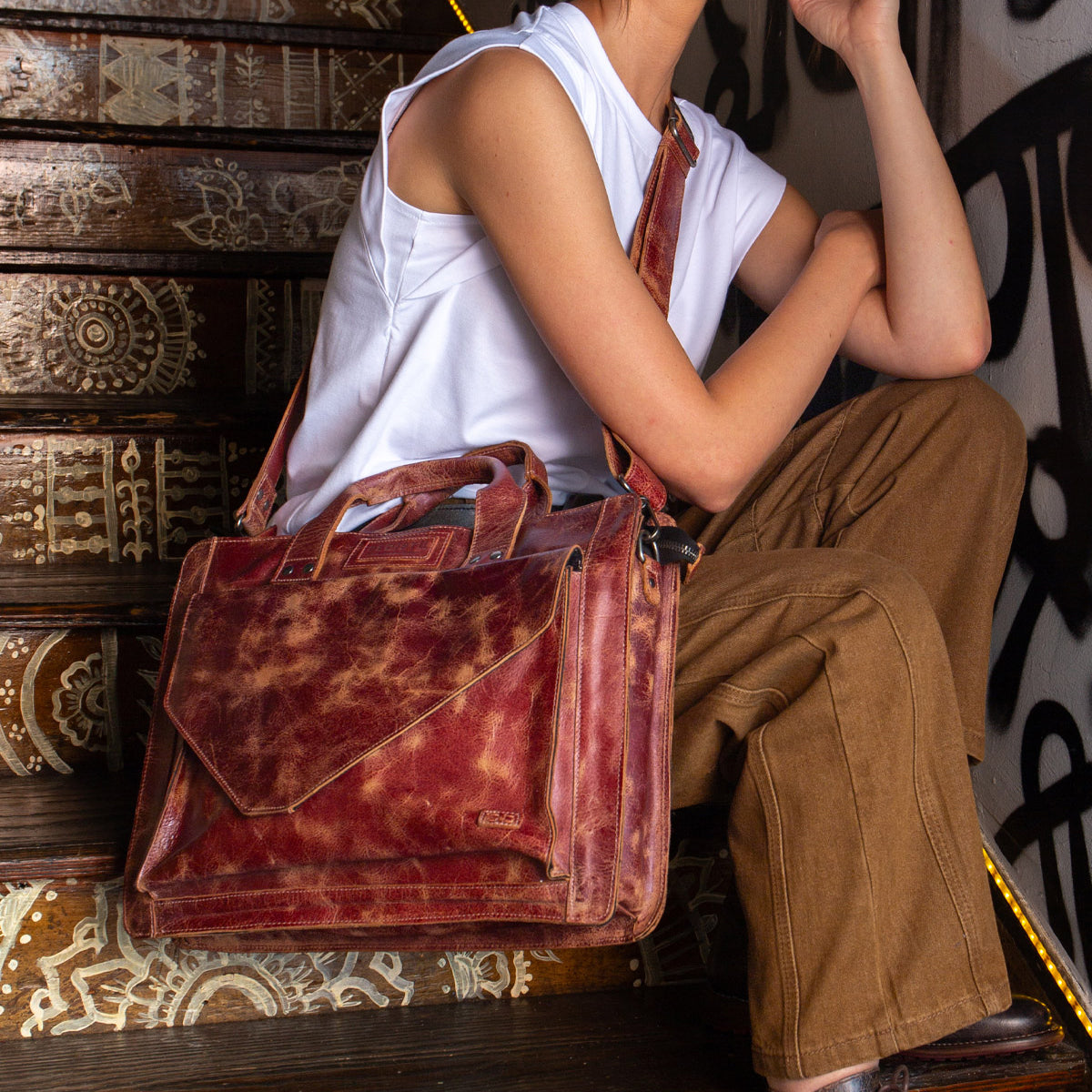 A person sits on patterned stairs, wearing a white sleeveless top and brown pants, holding the Bed Stu Depp distressed brown leather work bag that complements their minimalist style.