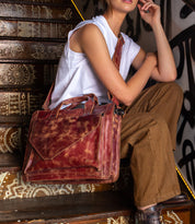 A person sits on patterned stairs, wearing a white sleeveless top and brown pants, holding the Bed Stu Depp distressed brown leather work bag that complements their minimalist style.