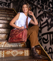 A woman sits on decorated wooden stairs in a white sleeveless top, brown pants, and boots, holding the Bed Stu "Depp" minimalist work bag. Behind her, bold black graffiti art covers the wall.