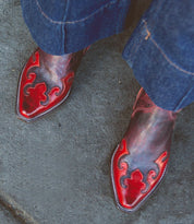 A pair of shiny red and black Deuce women's mid-calf cowboy boots by Bed Stu, featuring decorative stitching, worn with blue denim pants, showcasing a classic western style.