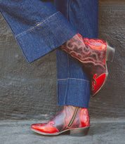 A person wearing blue denim jeans and Bed Stu's Deuce classic Western-style women's mid-calf cowboy boots is standing against a dark wall. The Deuce boots, made from vegetable-tanned leather, feature intricate red and black patterns with side zippers and pointed toes.