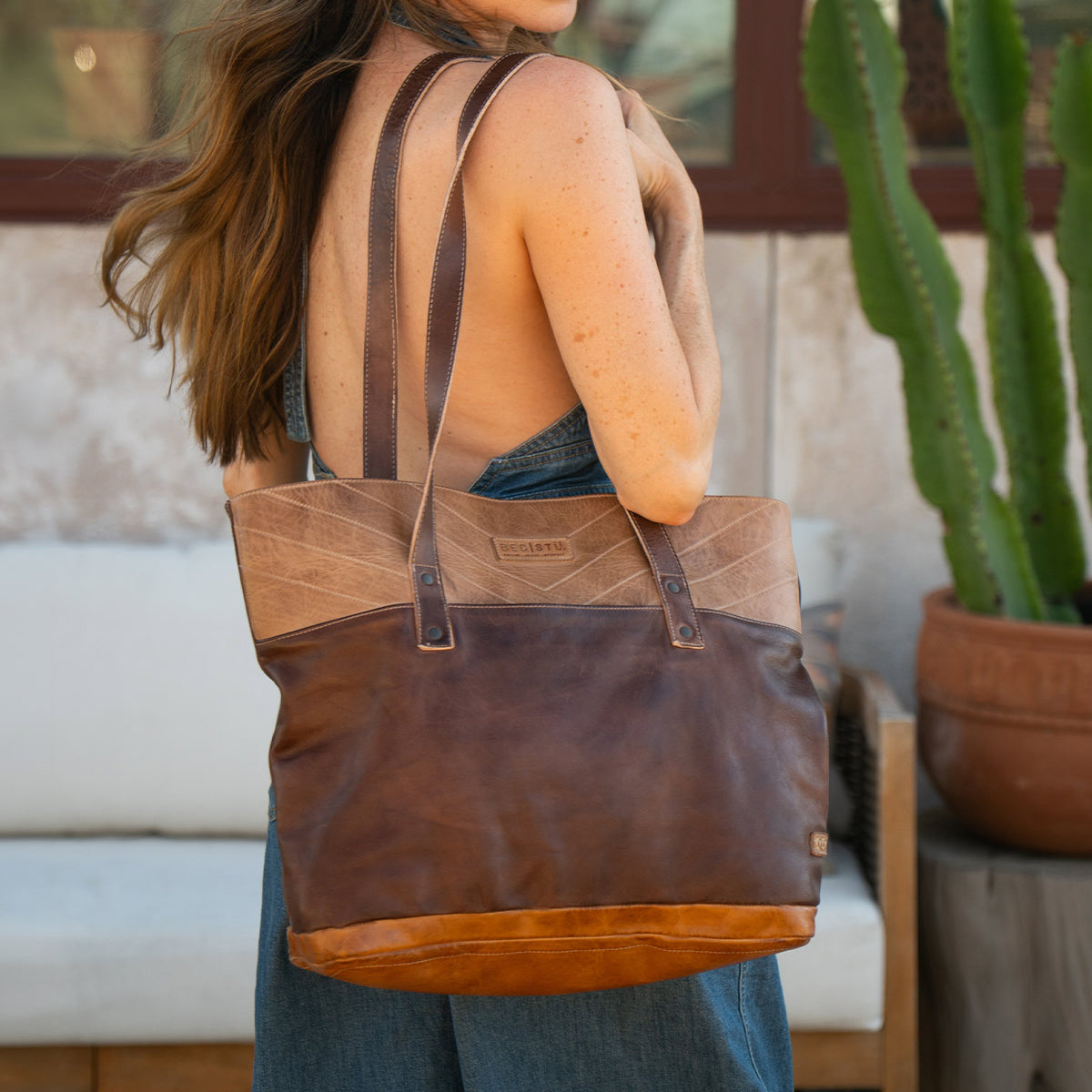 A woman with long hair wears a sleeveless jumpsuit and carries the Bed Stu Dome leather tote, featuring two-tone brown panels and a roomy interior—ideal for work essentials—while standing outdoors near a potted cactus and white sofa.