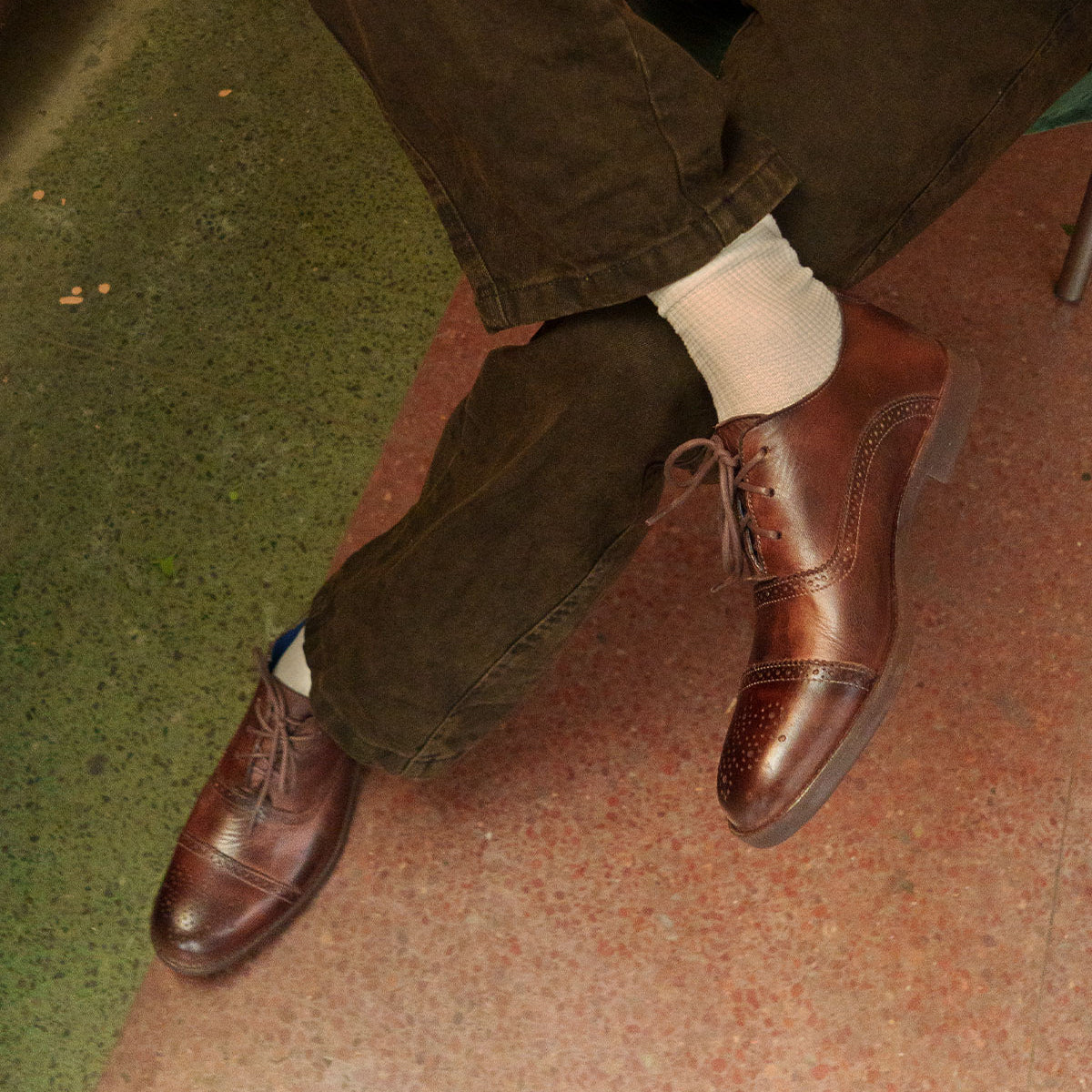 A person wearing Bed Stu's Donatello II versatile leather dress oxfords, paired with white socks and dark pants, is seated on a green and pink speckled floor.