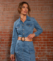 A woman stands before a brick wall wearing the Bed Stu Dreampath EM: a denim dress with long sleeves and a modern belt featuring an octagon metal buckle.