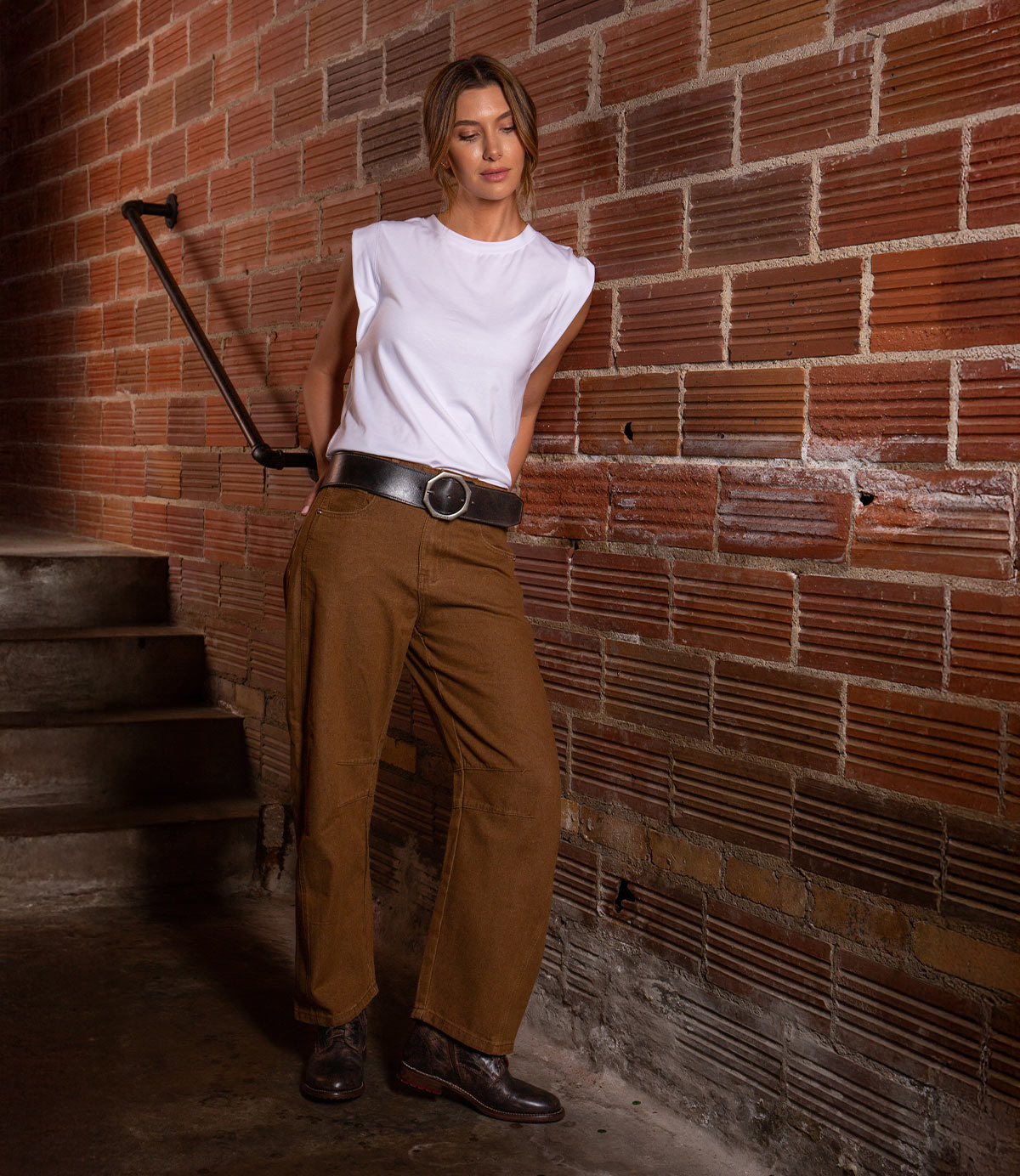 Wearing the Bed Stu "Dreampath" genuine leather belt with an octagon metal buckle, a woman in a white sleeveless shirt, brown pants, and black shoes stands by a brick wall near a staircase.