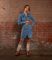 A woman stands before a brick wall wearing Bed Stu’s Dreampath denim dress, styled with an octagon-buckle leather belt and light brown cowboy boots.