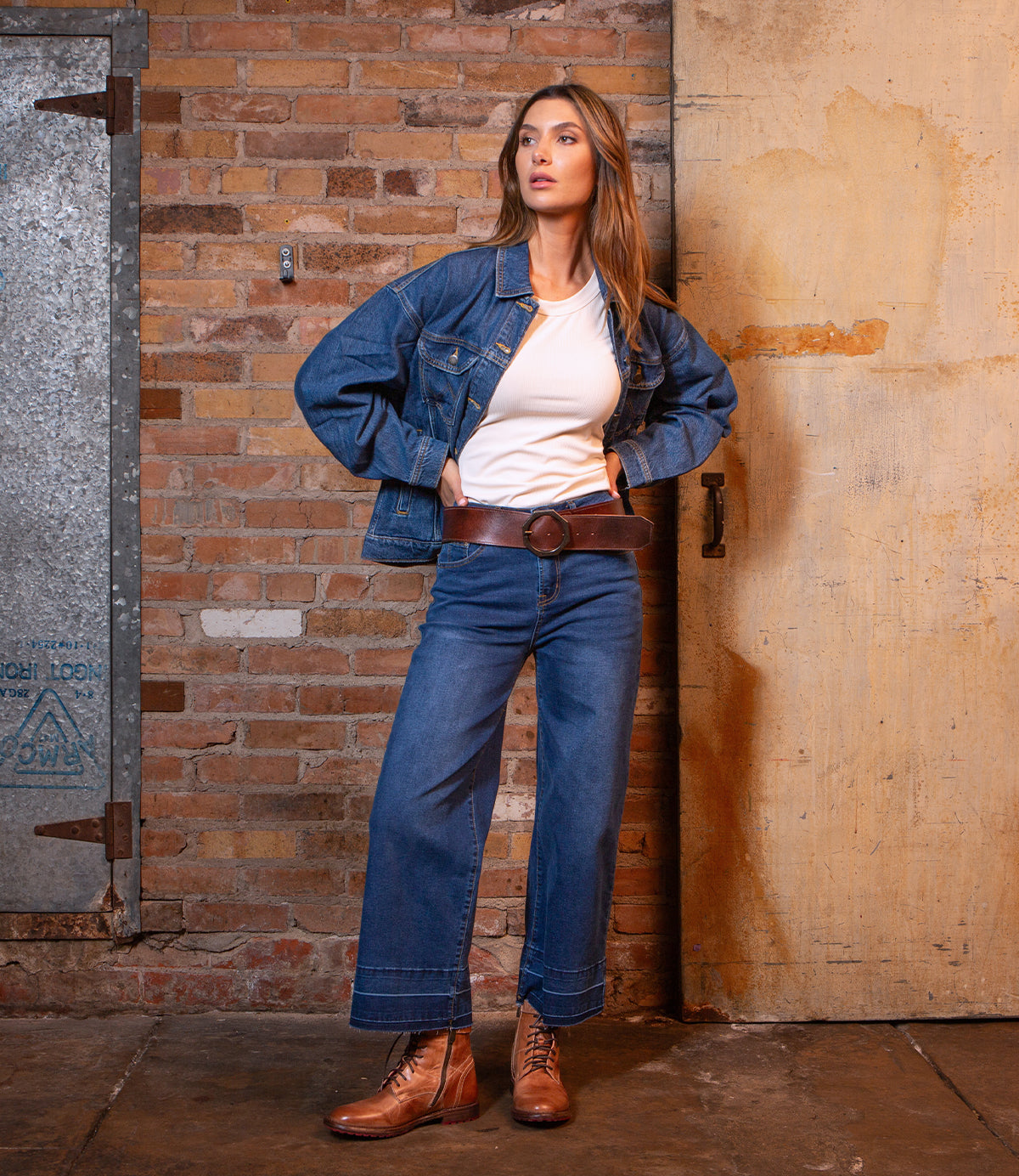 A woman stands against a brick wall wearing the Bed Stu Dreampath genuine leather belt with an octagon metal buckle, styled with a denim jacket, white shirt, wide-leg jeans, and lace-up boots.