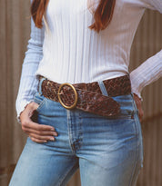 The individual is dressed in a white ribbed long-sleeve shirt, blue jeans, and the Bed Stu Dreamweaver belt, which boasts a wide brown woven design with vintage allure and a large brass-tone buckle.
