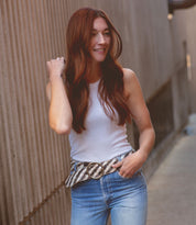 A person with long reddish-brown hair, wearing the Dreamweaver by Bed Stu—a white tank top and jeans complemented by a brass-tone buckle—stands in a narrow walkway. They smile slightly, one hand gently touching their hair, exuding a hint of vintage allure.