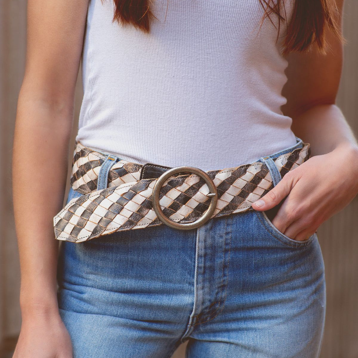 A person wearing a white tank top and blue jeans featuring the Dreamweaver from Bed Stu, a wide woven belt with vintage allure and a large brass-tone buckle.