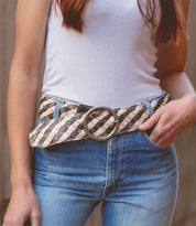 A person wearing a white tank top and blue jeans featuring the Dreamweaver from Bed Stu, a wide woven belt with vintage allure and a large brass-tone buckle.