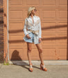 Woman in a white shirt and denim shorts, cinched at the waist with a Bed Stu Dreamweaver vintage belt featuring a round brass-tone buckle, posing in front of a garage door on a sunny day.