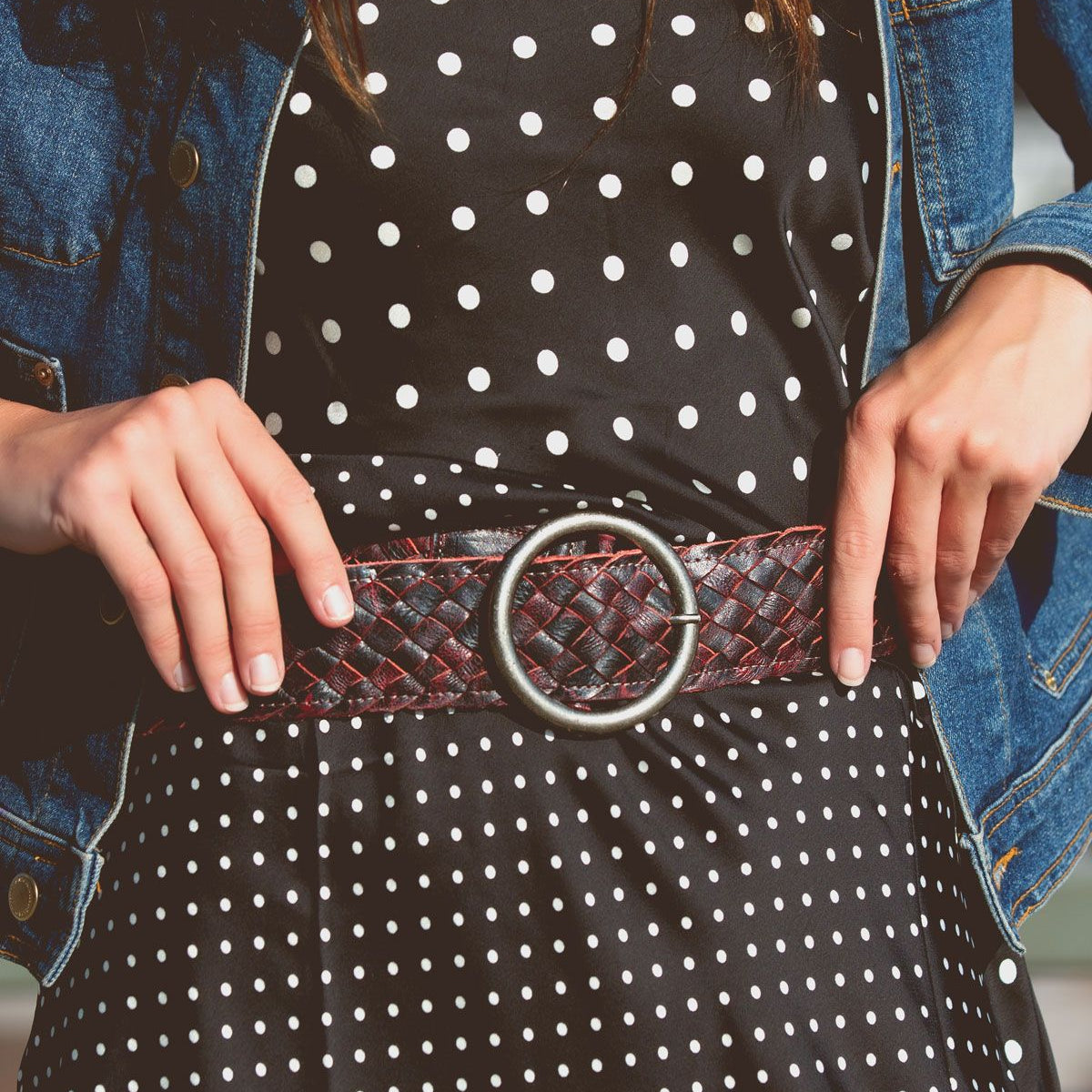 A person wears a black and white polka dot dress, the Bed Stu Dreamweaver wide brown woven belt with a brass-tone buckle, and a blue denim jacket.