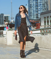 A woman in a black polka dot dress, denim jacket, and boots walks on a rooftop with city buildings in the background. She is wearing sunglasses and smiling, exuding vintage allure with her Bed Stu Dreamweaver distressed leather bag featuring a round brass-tone buckle.