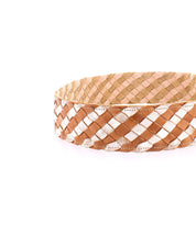 The Dreamweaver by Bed Stu is a woven basket with a checkered brown and beige pattern, featuring metallic accents and a brass-tone buckle, set against a plain white background.