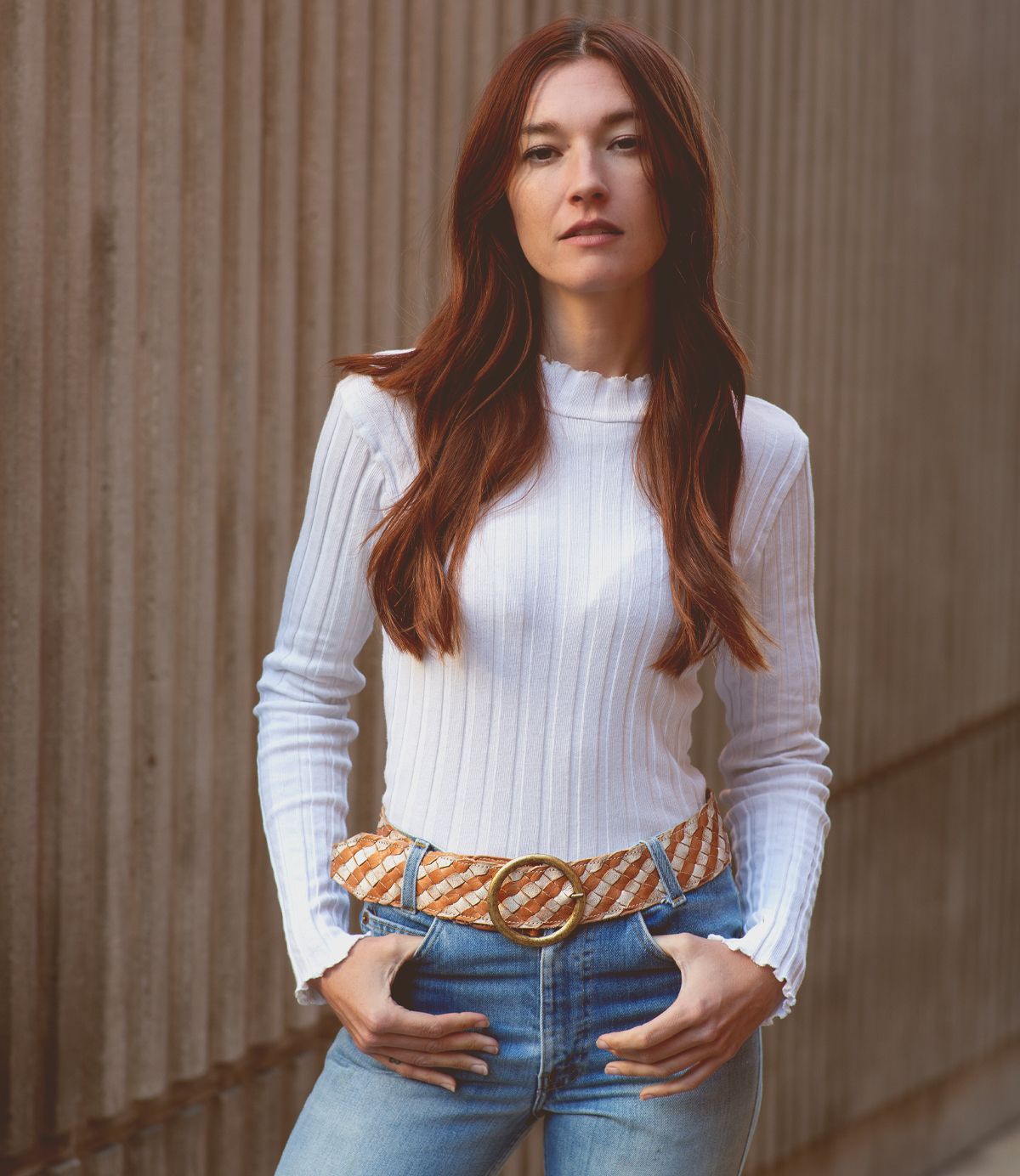 A woman with long hair leans against a textured wall, wearing the Dreamweaver white ribbed sweater by Bed Stu, paired with jeans and a patterned belt featuring a brass-tone buckle.