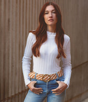 A woman with long hair leans against a textured wall, wearing the Dreamweaver white ribbed sweater by Bed Stu, paired with jeans and a patterned belt featuring a brass-tone buckle.