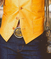 Close-up of a person wearing a Dreamweaver yellow leather vest with brown buttons, blue jeans with orange stitching, and a decorative round brass-tone buckle exuding vintage allure by Bed Stu. The person's hand holds a Bed Stu brown leather bag.