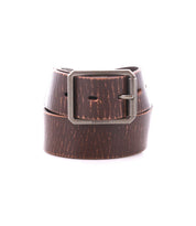 The Drifter belt by Bed|Stü, featuring a textured brown leather strap and a rectangular metal buckle, is rolled up against a plain white background, showcasing its minimal style.