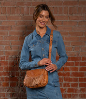 A woman in a denim dress stands by a brick wall, smiling and holding the Bed Stu Eastend—a stylish handmade bohemian leather purse with fringe accents.