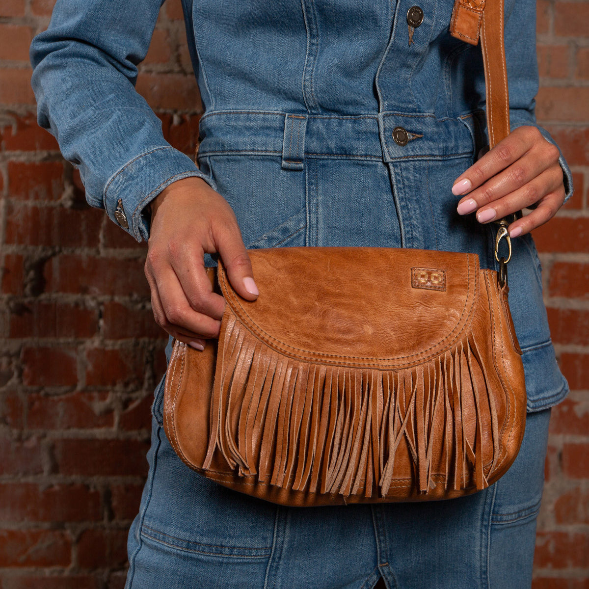 Person in a denim jumpsuit holds the Bed Stu Eastend bohemian leather purse with fringe details in front of a brick wall.