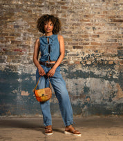 A woman stands by a distressed brick wall, wearing a sleeveless denim top, matching jeans, sandals, and carrying the Bed Stu Elect shoulder bag for a touch of vintage charm to her casual outfit.