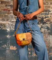 A person in denim stands against a weathered brick wall, holding Bed Stu's Elect—a chic shoulder bag with an adjustable strap and timeless vintage charm.