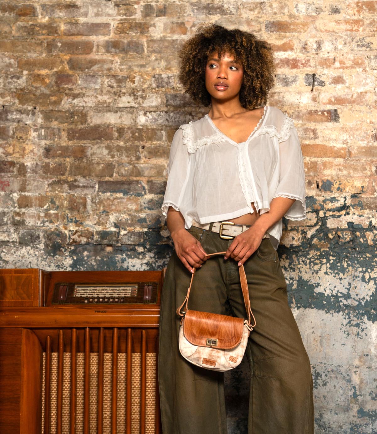 A person with curly hair stands against a brick wall, wearing a white blouse, olive pants, and holding the Bed Stu Elect shoulder bag with an adjustable strap in front of a vintage wooden radio.
