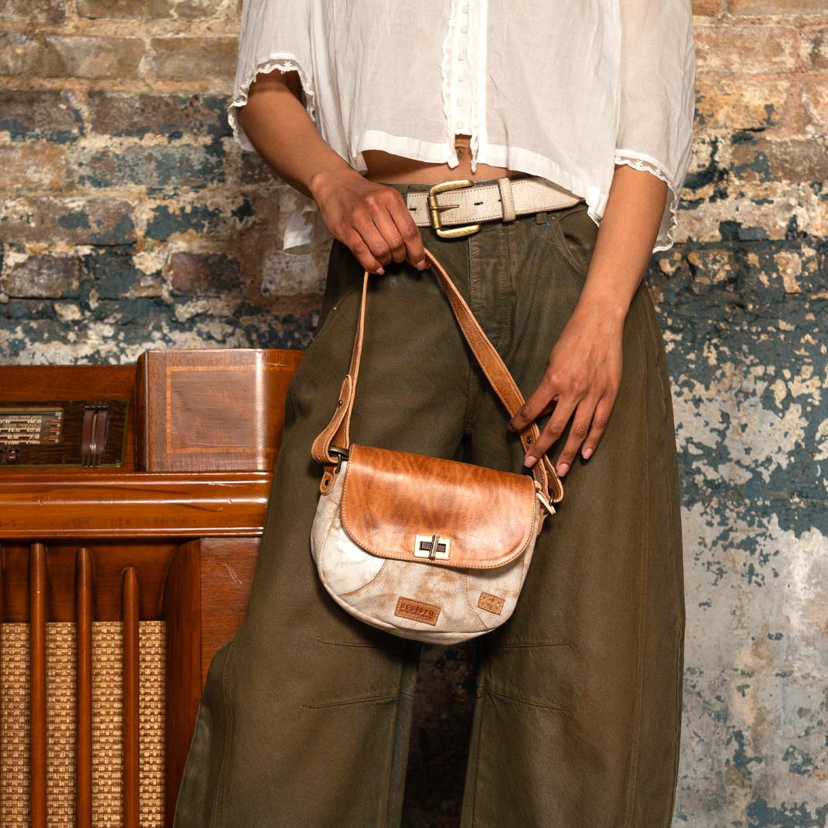 A person in a white blouse and green wide-leg pants stands by a worn brick wall, holding the Bed Stu Elect shoulder bag—a small brown and white bag with vintage charm, an adjustable strap, and buckle.