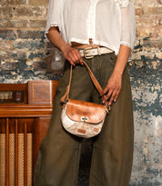 A person in a white blouse and green wide-leg pants stands by a worn brick wall, holding the Bed Stu Elect shoulder bag—a small brown and white bag with vintage charm, an adjustable strap, and buckle.