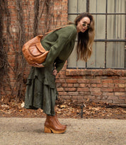 A woman showcases her fashion self-expression on a city sidewalk, leaning forward in an olive green outfit and brown boots, wearing sunglasses and carrying the large brown Bed Stu Ellasphin bag over her shoulder.