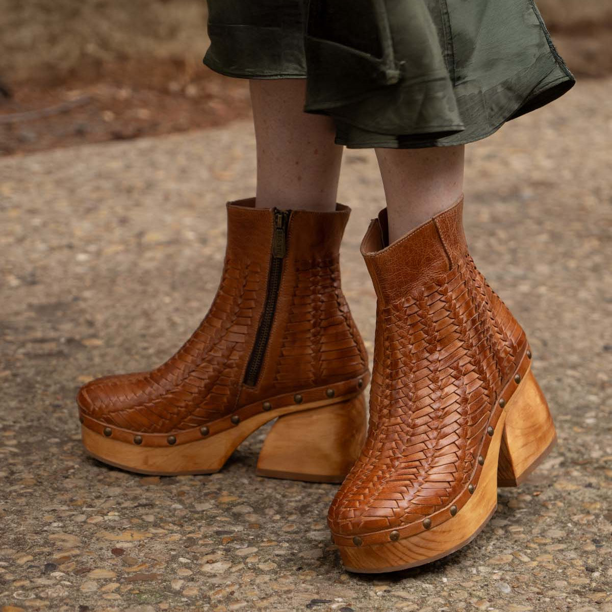 A person wearing Bed Stu's Ellasphin brown handwoven wooden platform boots with a visible zipper and sculptural style stands on a gravel path, showcasing fashion self-expression.