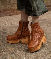 A person wearing Bed Stu's Ellasphin brown handwoven wooden platform boots with a visible zipper and sculptural style stands on a gravel path, showcasing fashion self-expression.