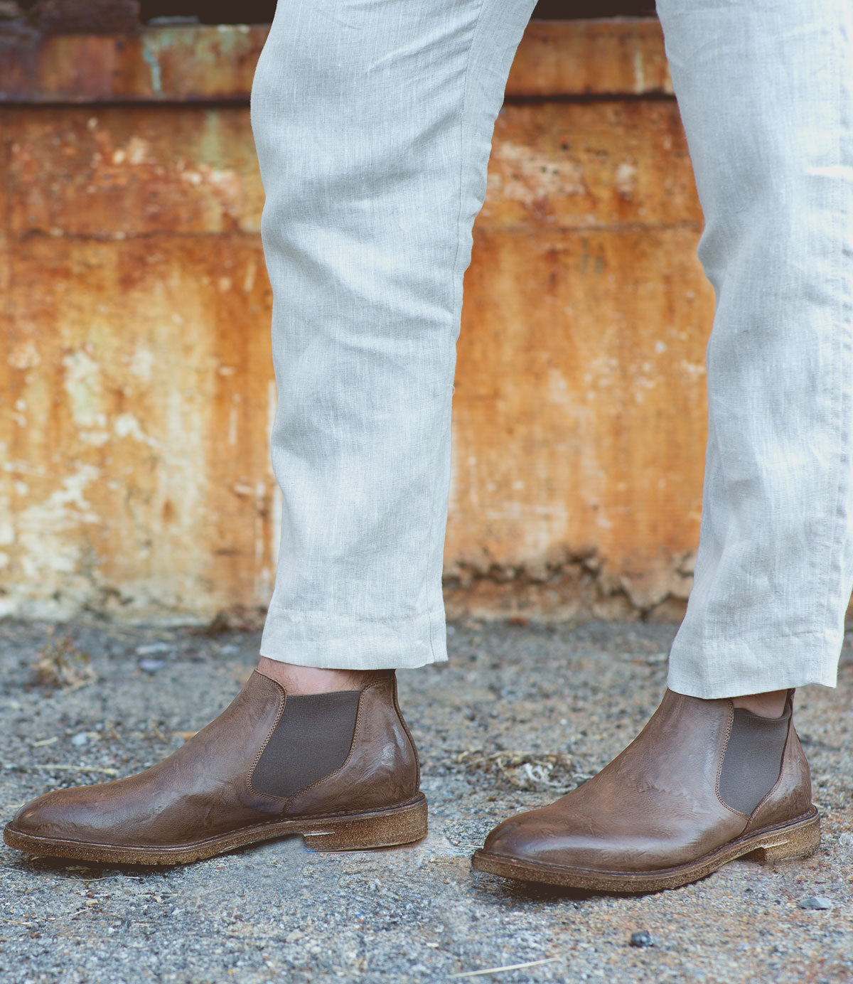Person wearing light-colored pants and the Elude men's leather boots by Bed Stu, handcrafted in Italy, standing on gravel with a rusted metal wall in the background.