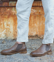 Person wearing light-colored pants and the Elude men's leather boots by Bed Stu, handcrafted in Italy, standing on gravel with a rusted metal wall in the background.
