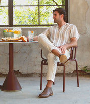 A man in a striped shirt and beige pants, complemented by Elude men's leather boots handcrafted by Bed Stu in Italy, sits by a small table with drinks and fruit. He is looking to the side in a sunlit room with an old, textured wall and a window showing greenery outside.