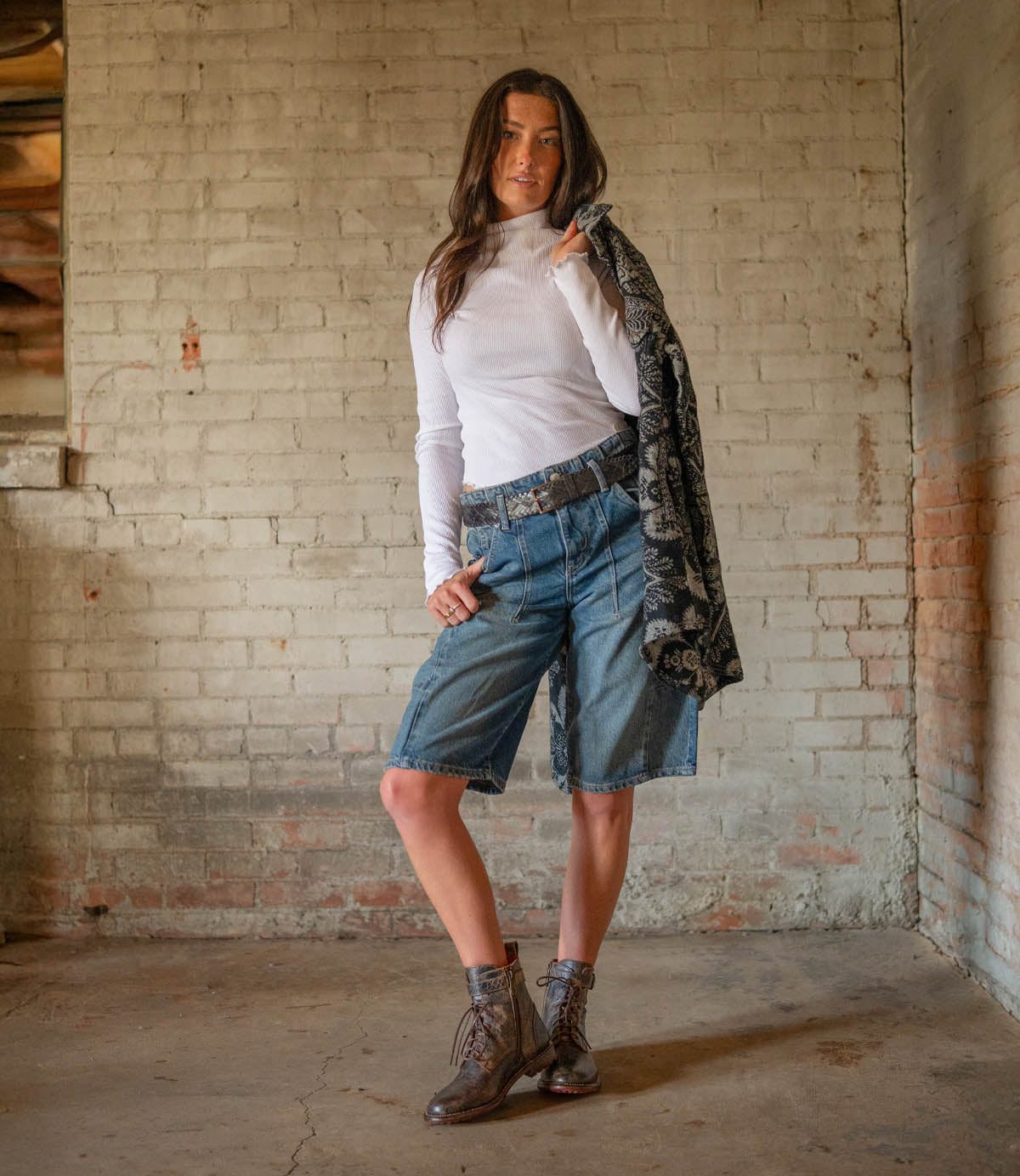 A woman stands before a brick wall wearing a white turtleneck, denim shorts, and Bed Stu’s Endure lace-up ankle boots, holding a dark patterned jacket over her shoulder.