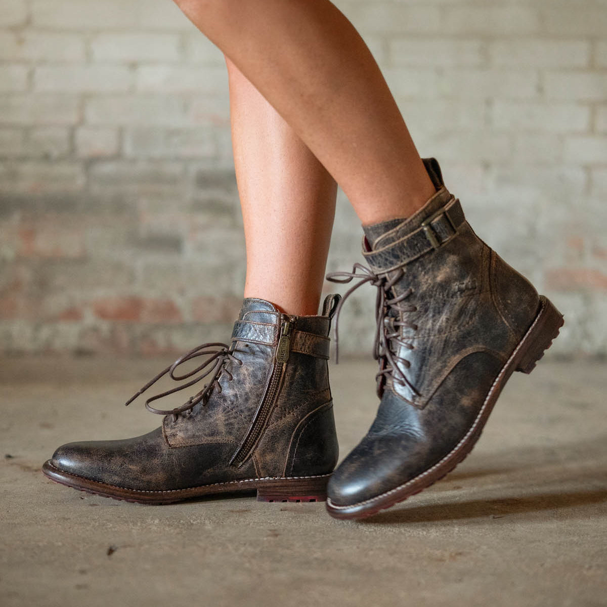 A person stands on a concrete floor before a brick wall, wearing Bed Stu’s Endure: distressed brown lace-up ankle boots with buckles and leather lining, perfectly channeling rugged sophistication.