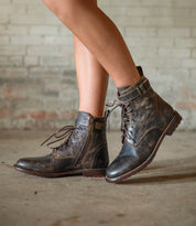 A person stands on a concrete floor before a brick wall, wearing Bed Stu’s Endure: distressed brown lace-up ankle boots with buckles and leather lining, perfectly channeling rugged sophistication.