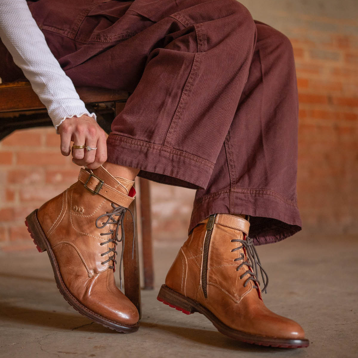 A person wearing Bed Stu's Endure rugged lace-up ankle boots with leather lining, brown pants, and a white long-sleeve top sits on a wooden chair against a brick wall, showcasing effortless rugged sophistication.